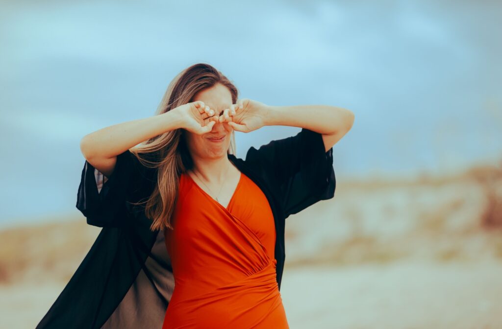 A young woman in a red dress rubs her eyes in pain from the sand and wind hitting her eyes while on the beach