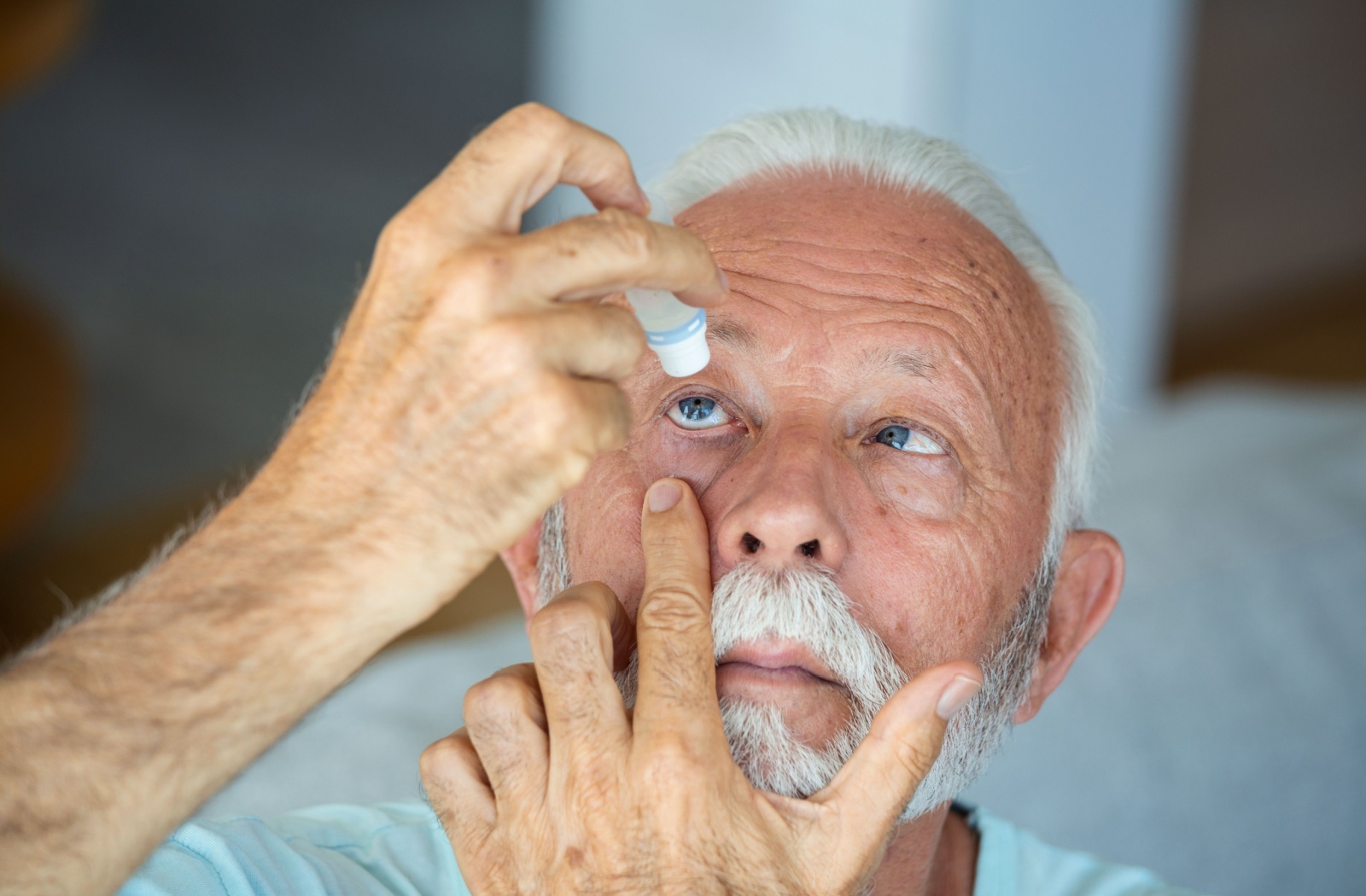 An older adult applies eye drops into their right eye