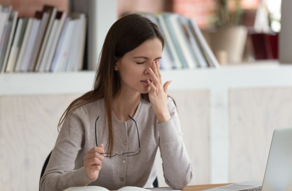 A young adult removes their glasses to rub their left eye due to experiencing eye strain from their computer.