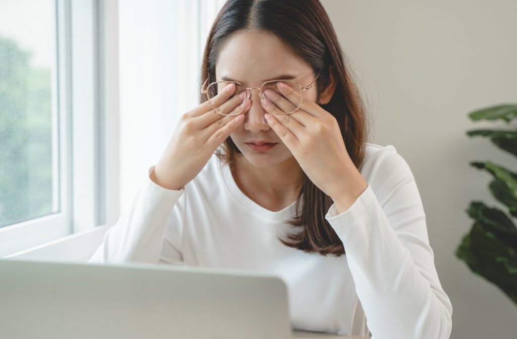 A person working at their computer, pushing their glasses out of the way and rubbing their eyes due to discomfort when blinking.